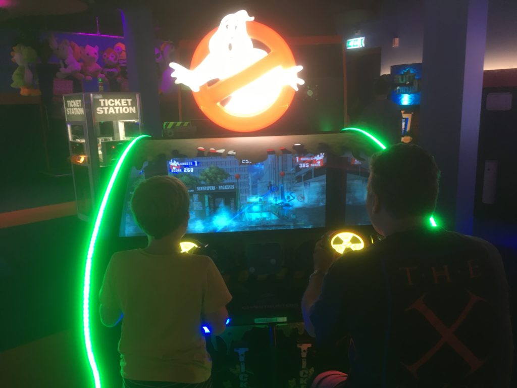 Paul and Yuri (on the left) are sitting in a machine/capsule with green lights on the side. Above is the Ghostbusters sign. In front of them is the screen. 