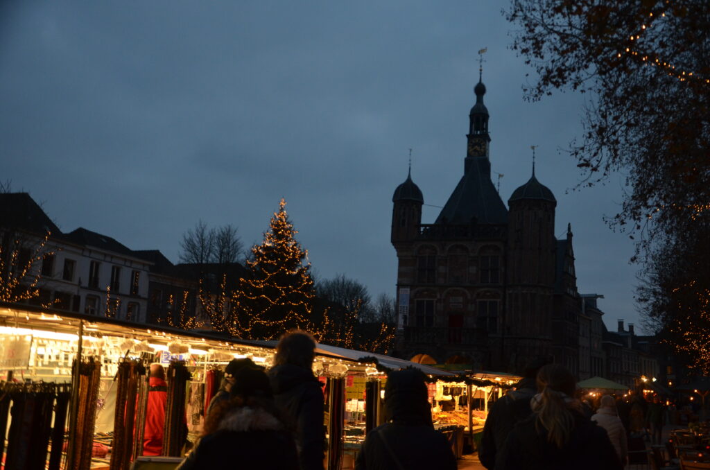 Christmas market on the Brink. In the evening, lighted up stalls and a a Christmas tree in the middle. In the forefront people walking.