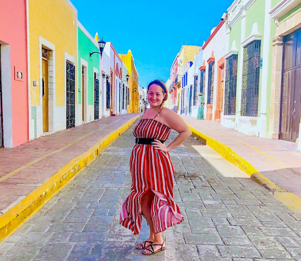 Shelley from Travel Mexico Solo. Shelley stands in the middle of a street with colored houses on both sides. She has a red dress on.