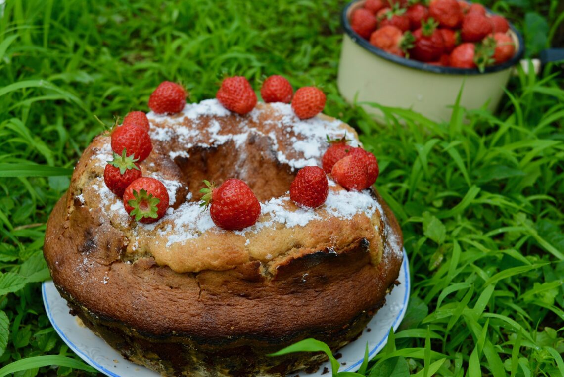 Keks, a sort of bundt cake topped with straweberries on a plate standing in the grass, further back a bowl with strawberries