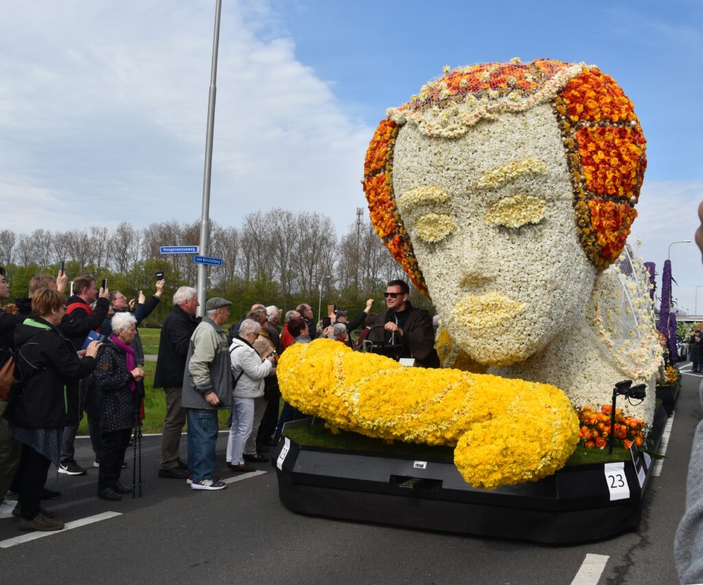 A flower float during the Bloemencorso. A car with a sculpture made with flowers on it. The sculture is a woman's face. White, yellow and orange flowers are used to sculpt the face. People are standing on the left side of the parade. A blue sky with some clouds above. 