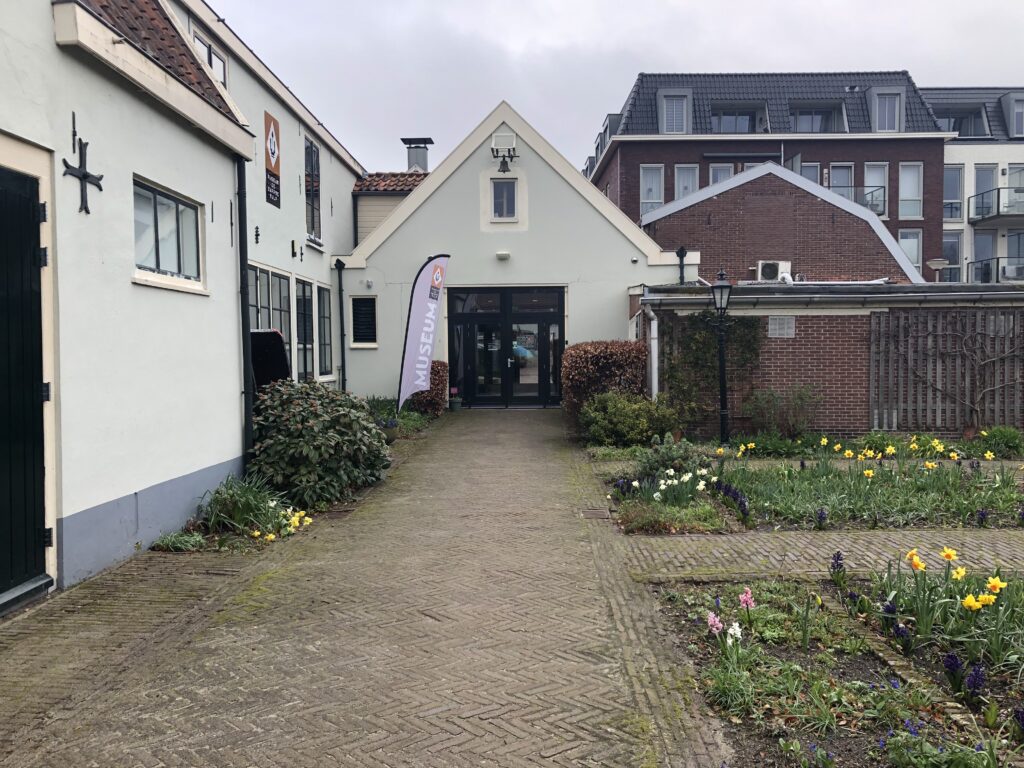 The entrance to Museum the Black Tulip. You see a brick path leading to a small white building. The doors are black and function as the entrance to the museum. On the right side of the path are square flower beds. On the left side are walls from the adjacent buildings. There's an overcast sky. 