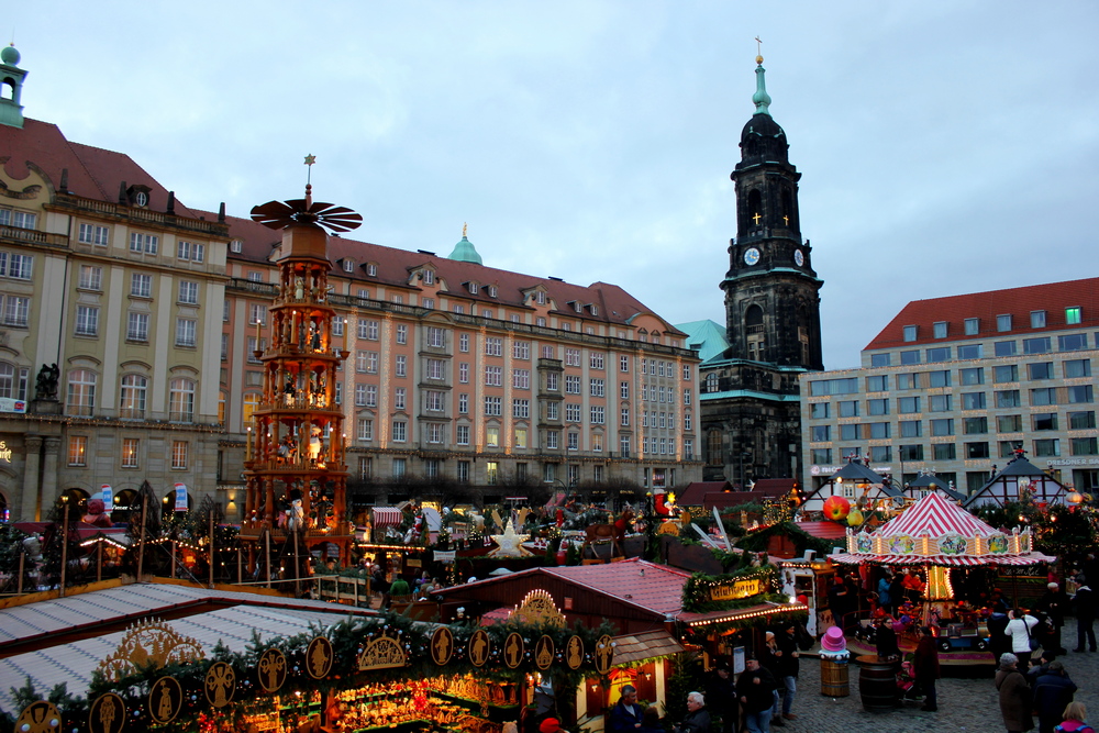 Dresden Christmas market by Berlin Travel Tips, a Christmas market on a square. With large buildings behind it and the Frauenkirche. A large Christmas display to the left in the market