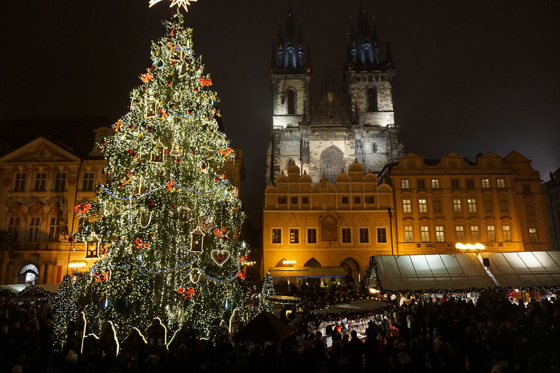 Prague Christmas markets by Andoreia, in the dark. An large illuminated Christmas tree to the left and a large lit up building to the left with a Christmas market in front of it