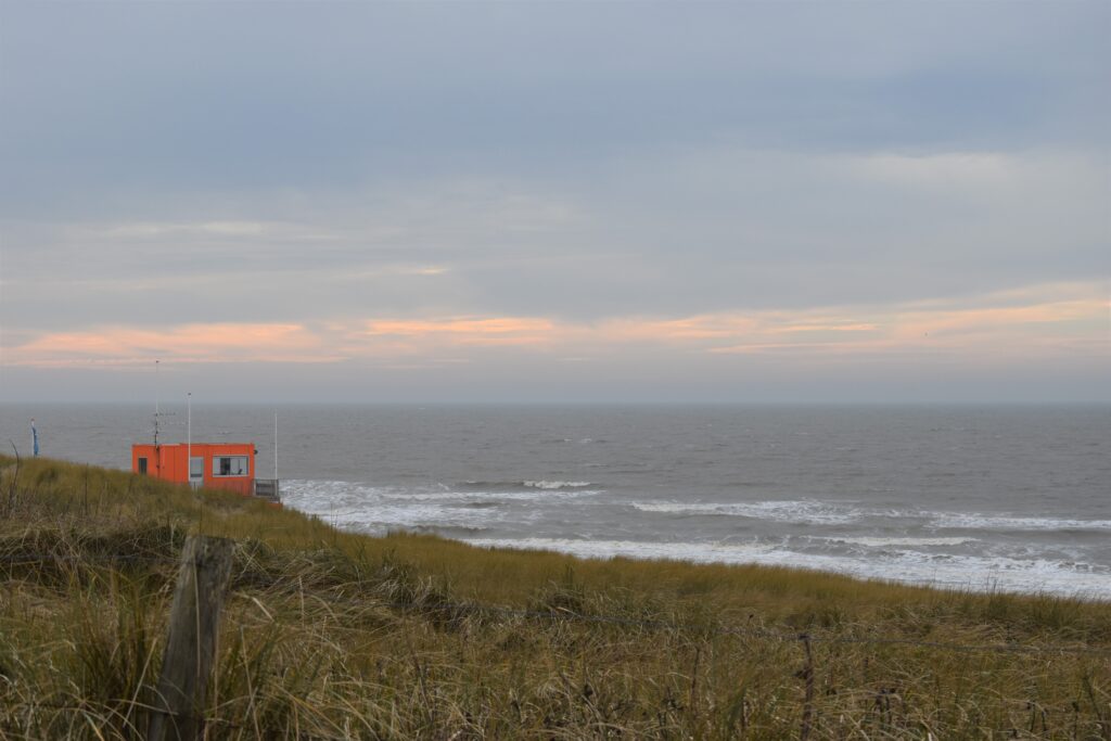 Another beautiful sea view, the grass on the dunes below, above it the sea and above that the clowded sky, on the left a orange building sticking out above the dunes