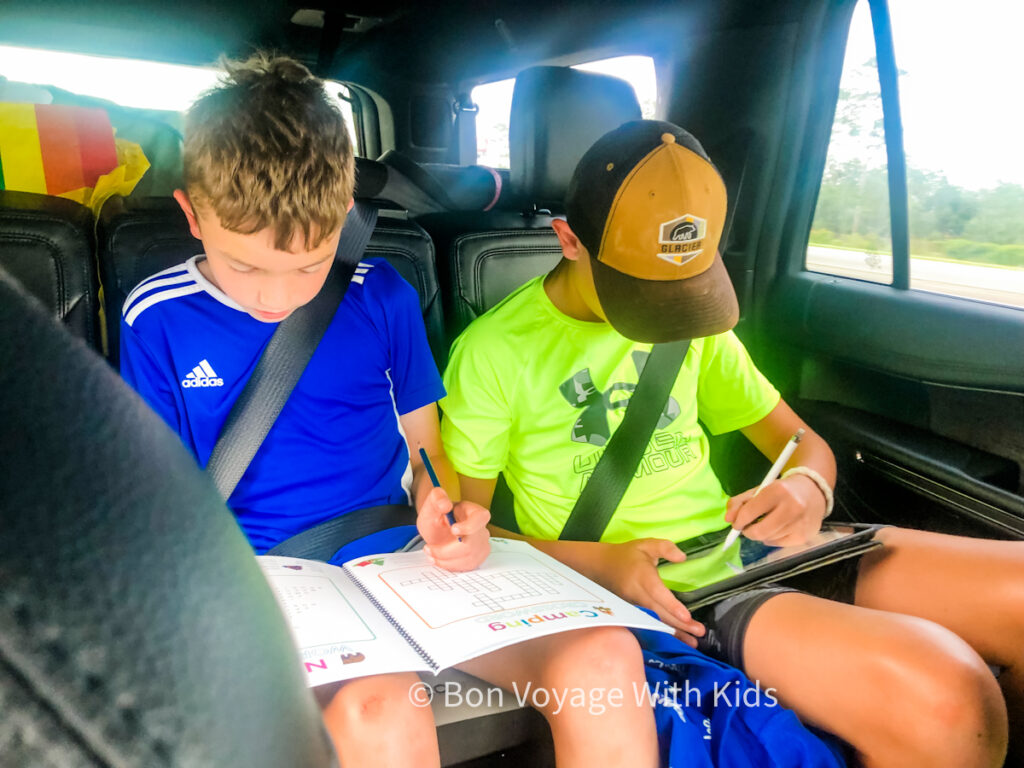 Listening to podcasts in the car by Bon Voyage With Kids, two boys sitting in the backseat, one with a tablet and a head on, the other with a book