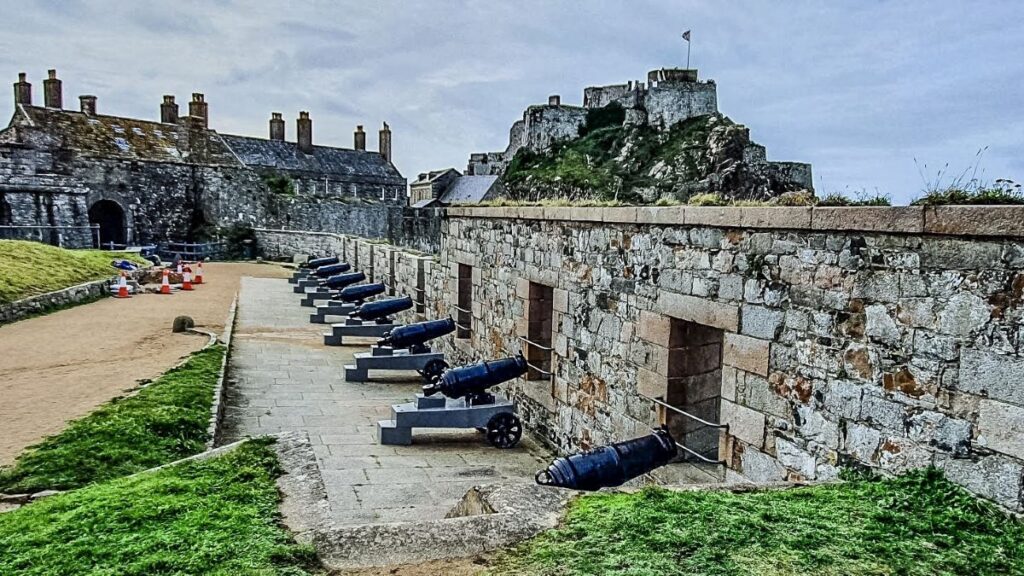 Cannon at Elizabeth Castle Jersey by Grey Globetrotters, a row of canons in the middle right, against a wall where holes are in. Behind a building and further back the ruin