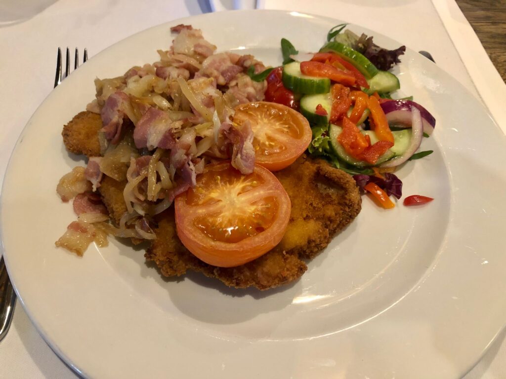 Farmer's schnitzel on a white round plate served topped with fried bacon and onions, tomato and some salad on the side