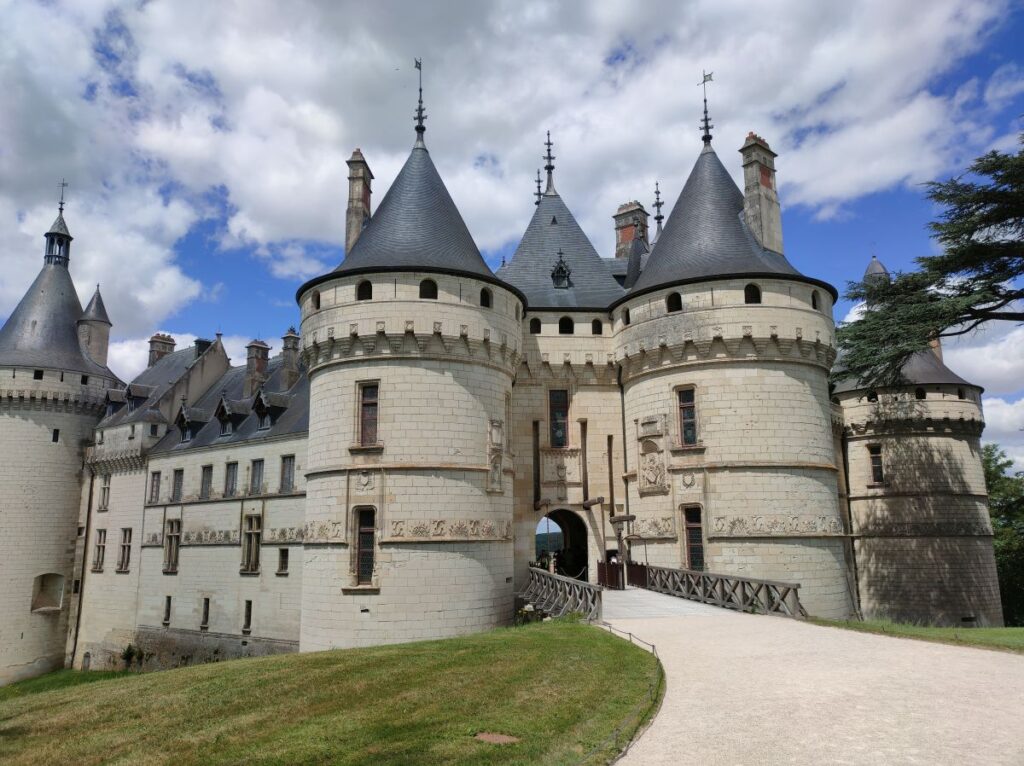Chateau de Chaumont by Ipanematravels, a pathruns towards the entrance to the castle. A grass field on the left side. A grey colored walls, with blue tiled roofs. Turrets in front at the entrance