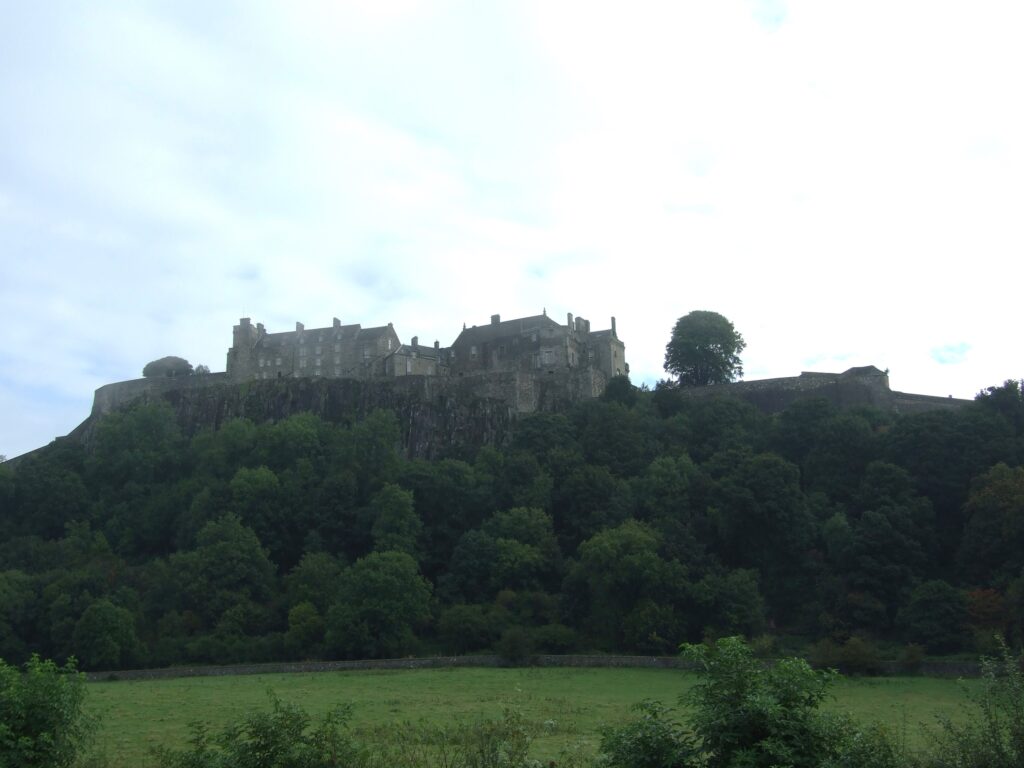 Stirling Castle, as seen from a bit further away. It perches on top of a rock. Trees and grass in front of it